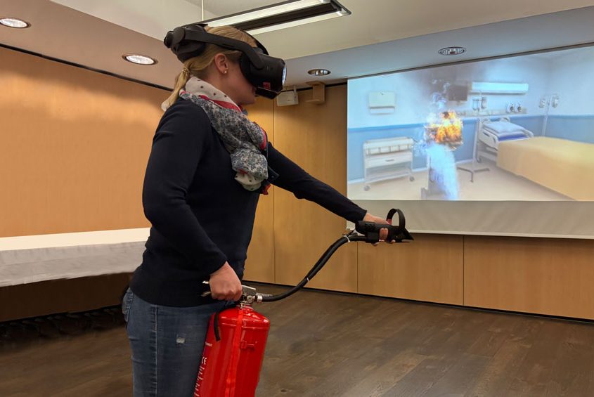 Virtuelles Löschtraining -Frau mit VR-Brille und Löschattrappe. Auf der Leinwand sieht man den virtuellen Brand der gelöscht wird
