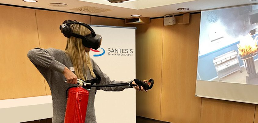 Frau beim virtuellen Löschtraining - sie hält die Feuerlöscher-Atrappe und auf der Leinwand kann man die virtuelle Umgebung miterleben.
